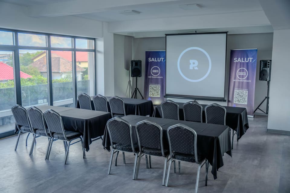 Sala aranjată în format de curs: mese acoperite cu fețe de masă negre, așezate în rânduri, cu scaune orientate spre ecran.